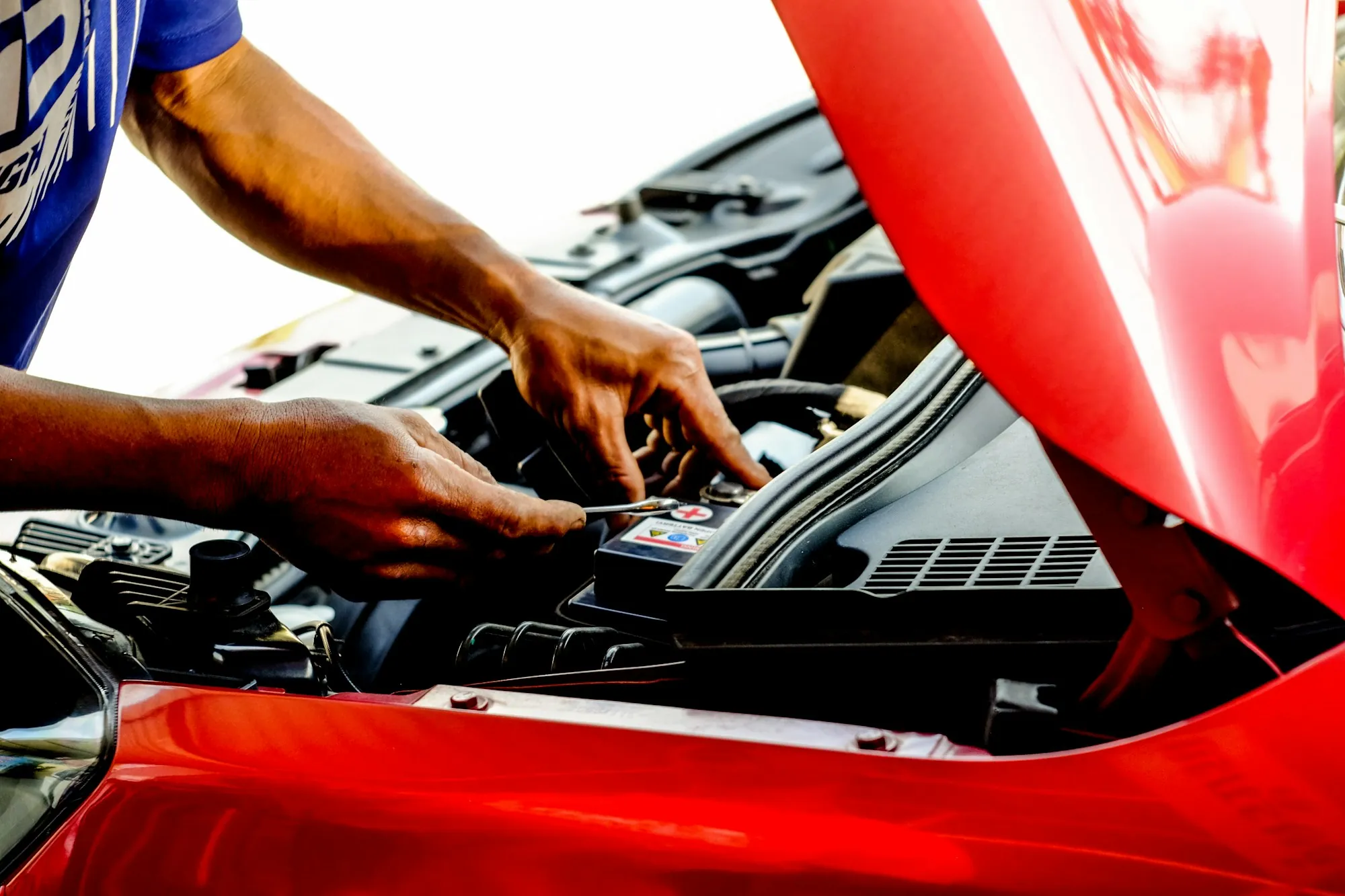 Hands working on the engine of a red car with the hood up